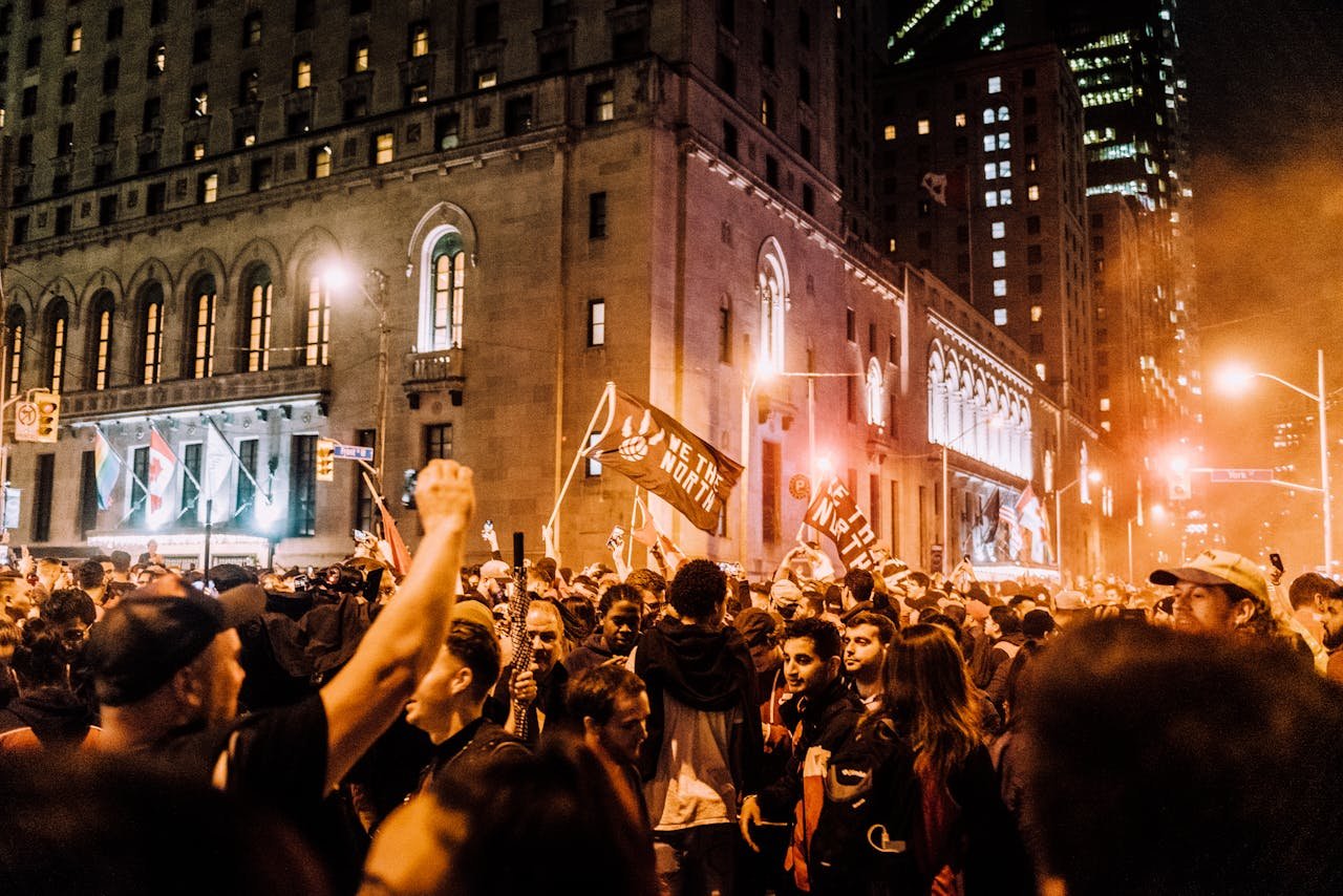 Vibrant city street scene at night featuring a large crowd with flags, celebrating under illuminated buildings.