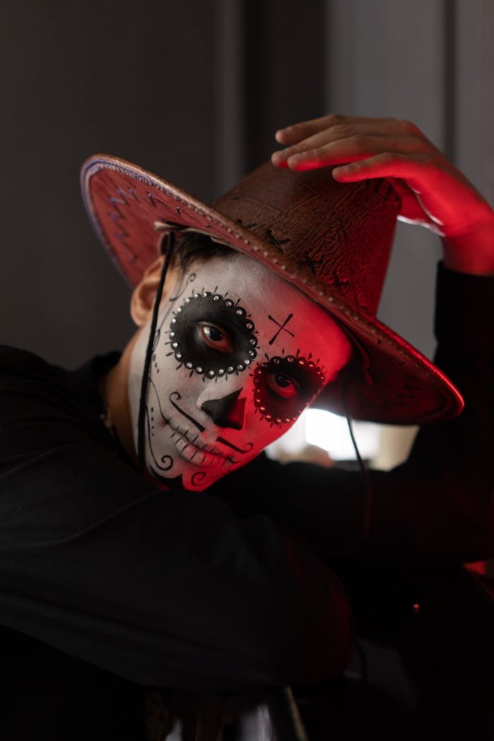 A dramatic portrait of a person wearing intricate Halloween face paint and a cowboy hat, lit with moody red lighting.